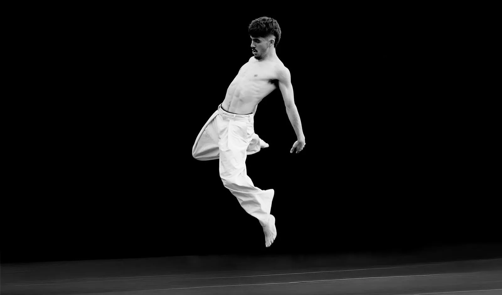 PierreDo en plein saut, torse nu et pantalon blanc, photo en noir et blanc sur fond noir